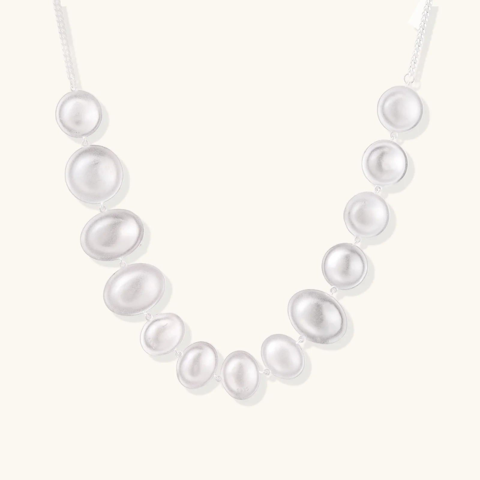 Graduated pearl-finish silver discs forming an organic bubble bib necklace on delicate chain against cream background.